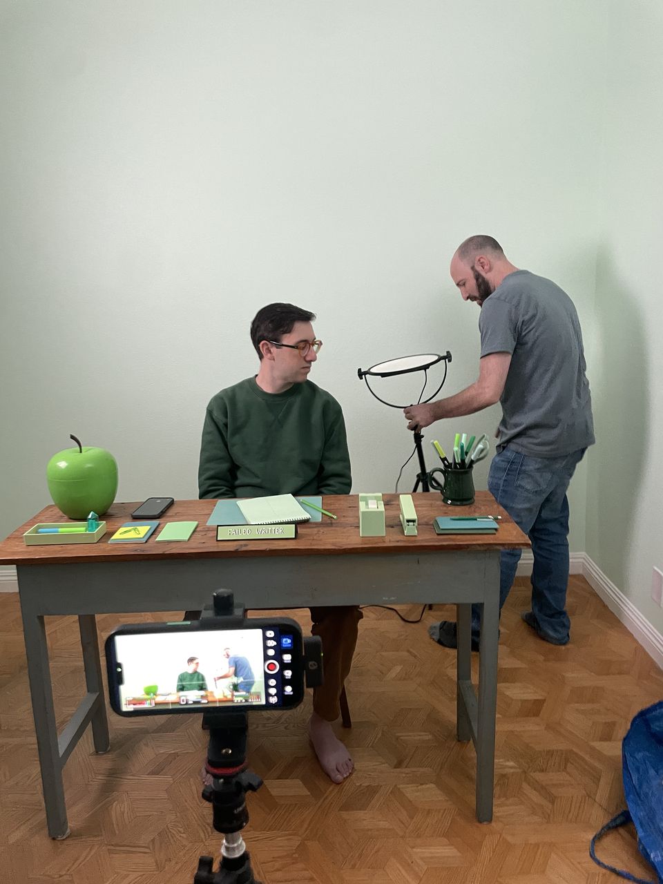Justin Michael at a desk on his set while a collaborator adjusts a ring light, a phone on a tripod in the foreground